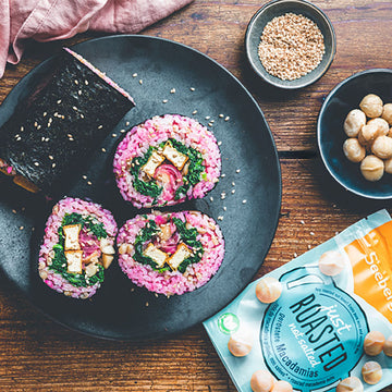 Sushi mit pinkem Reis liegt auf einem Teller, umgeben von Schalen mit Nüssen.